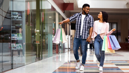 Black Friday Sales. Happy African American Couple Walking With Shopping Bags In Mall, Looking And Pointing At Showcases, Young Spouses Making Purchases Together In Modern Department Store, Copy Space