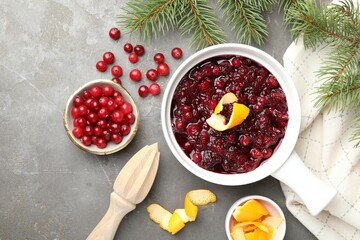 Tasty cranberry sauce in gravy boat, berries, orange peels, squeezer and fir branches on grey table, flat lay