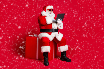 Full length photo of retired old man white beard sit giftbox hold laptop take orders online wear x-mas santa costume glove coat belt sunglass cap boot isolated red color background