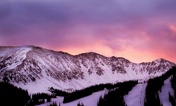 A vibrant winter sunset paints the snowy mountains in shades of pink and purple. 