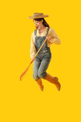 Female farmer with rake jumping on yellow background