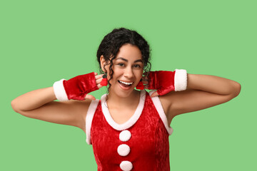 Beautiful young African-American woman in festive earrings and Santa Claus costume on green background. Christmas celebration