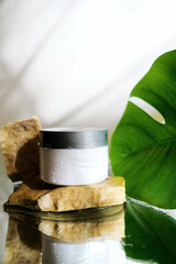 White jar with skincare product. Mirror reflection wet surface. Tropical nature. Wellness, mockup and beauty concept.