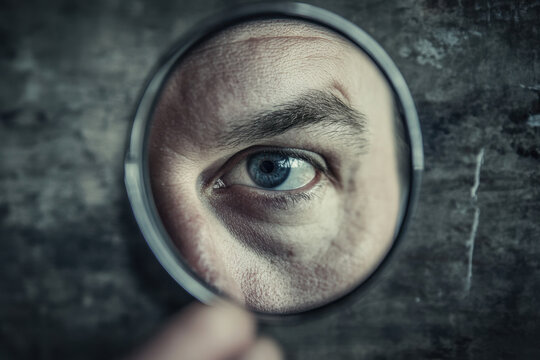 Close-up of a man's blue eye peering through a magnifying glass against a textured dark background, creating a mysterious and intriguing atmosphere.