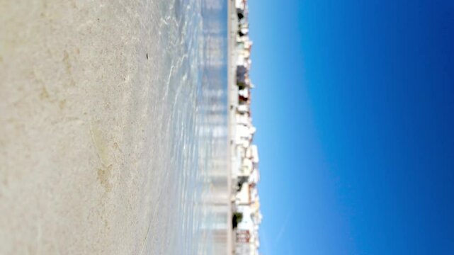A picturesque coastal town with whitewashed buildings reflects on the calm waters of a lagoon, under a vibrant blue sky.