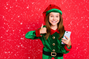 Portrait of cheerful schoolkid with long red head haircut showing thumb up holding gadget wearing...