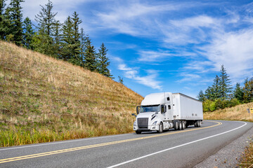 Popular comfortable white big rig semi truck transporting cargo in dry van semi trailer running on the winding mountain road between two hills