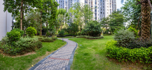 Panoramic of a courtyard garden or communal green outdoor space in a modern residential apartment building complex, featuring a well-manicured lawn, lush greenery, and a winding brick pathway