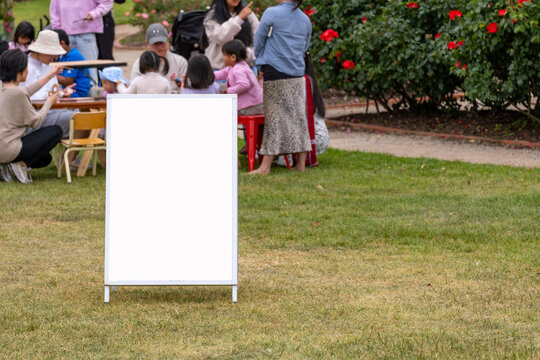 Background texture of a blank white information sandwich board mockup or A-frame sign standing on grass with a blurry view of family entertainment event or outdoor social gathering activity.