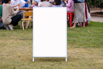 Background texture of a blank white information sandwich board mockup or A-frame sign standing on grass with a blurry view of family entertainment event or outdoor social gathering activity.