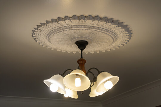 Classic outdated ceiling light fixture, adorned with ornate vintage Victorian-era decorative elements, is illuminated against a ceiling rose medallion with intricate plasterwork with a circular shape