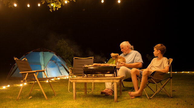 Grandfather seated on a chair by a small table children, holding little dog and barbecue grill, enjoying a meal or snack together indicating a night of camping joys of outdoor adventures concept.