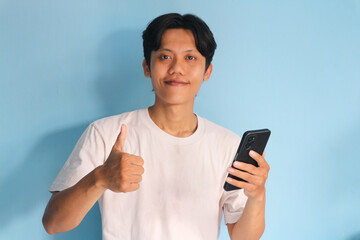 Young Asian man holding his mobile phone with happy expression and thumb up gesture	