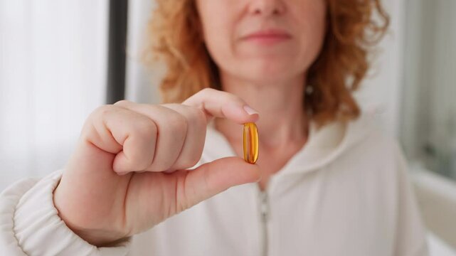 Close-up woman&rsquo;s hand shows orange capsule Omega D3 in camera and puts it near her mouth white background smily Health, complex vitamins people. Taking medication, pills. Vitamins for women's health