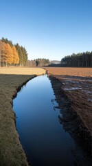 A serene landscape featuring a winding stream beside fields and trees under a clear sky.
