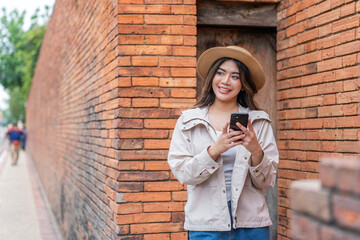 Asian Tourist Woman In Hat and Jacket Using Phone Beside A Brick Wall in Chiang Mai, Thailand