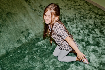 Cheerful little girl in cotton shirt and grey leggins sitting on the floor next to soft green slide after rolling down during play