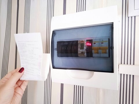 The hand holds the receipt in front of the electricity meter at home. Monthly electric bill. Payment for utilities. Tax increases. Energy saving.