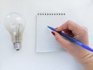 An incandescent lamp is lying on a white table, a woman's hand is writing with a pen in a notebook. Payment for utilities . The bill for electricity consumption.