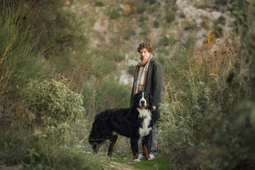 A woman walks with her Bernese Mountain Dog through a dense forest, surrounded by greenery.