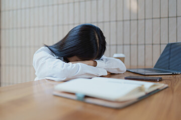 Young woman resting her head on arms at a wooden table in a cozy cafe, expressing fatigue and stress, with coffee cup and notebook nearby enhancing the scene's atmosphere