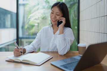 Young woman in a bright office talking on the phone while taking notes, with a laptop and coffee cup on a wooden desk, showcasing productivity and professionalism.