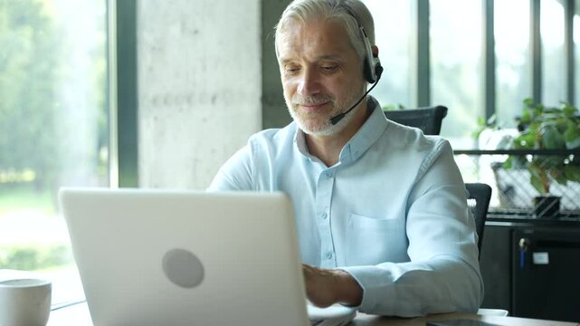 Smiling businessman using headset when talking to customer. Tech support manager in headset consulting a client. Happy mature male customer support executive working in office.