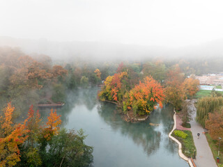 A misty autumn lake with vibrant orange, red, and green foliage surrounds the calm water. The fog adds a serene and mysterious atmosphere to the scene, with trees reflected on the still lake surface