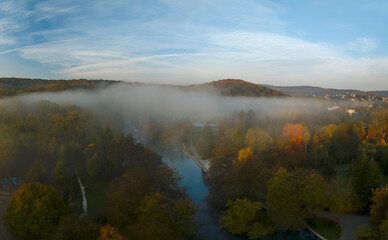A misty autumn lake with vibrant orange, red, and green foliage surrounds the calm water. The fog adds a serene and mysterious atmosphere to the scene, with trees reflected on the still lake surface