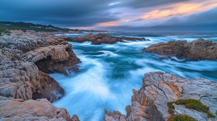 Fototapeta premium A stunning view of the crashing waves against the rugged coastline during a dramatic sunset. The blue water is smooth and flowing, while the sky is a mix of dark grey and orange.