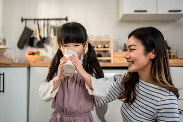 Asian little daughter drinking a glass of milk with mother in kitchen.