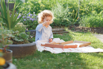 Little curly pretty girl sitting on lawn and eating delivered pizza from box in summer shiny...