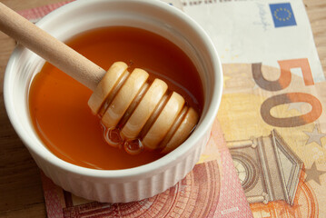 A bowl of organic honey with a honey spoon on Euro banknotes, symbolizing honey prices and bee value.