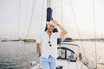 A cheerful man wearing a hat and casual attire stands on a sailboat. He's enjoying a sunny day at...