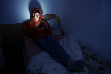 Teenager using a smartphone lying in bed late at night, playing games, watching videos online, and...