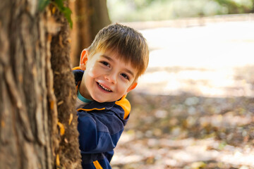 A little boy peeking out from behind the tree, smiling happily. Portrait of cute joyful child spending time in city park. Child emotions concept. Copy ad text space