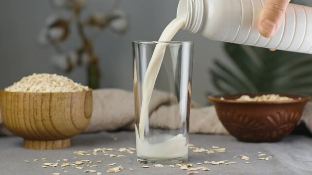 A hand is spilling oat flakes into a glass of natural oat milk on the table. Alternative vegetarian drink for a healthy diet. Slow motion