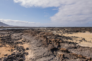 White sand beach with solidified volcanic lava flows, black rocks. Mountains in the background and sky with big white clouds. Caletón Blanco, Lanzarote, Canary Islands, Spain