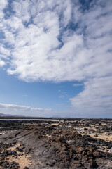 White sand beach with solidified volcanic lava flows, black rocks. Mountains in the background and sky with big white clouds. Caletón Blanco, Lanzarote, Canary Islands, Spain