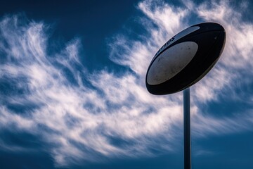 A rugby ball is mounted on a pole against a blue sky with white clouds.