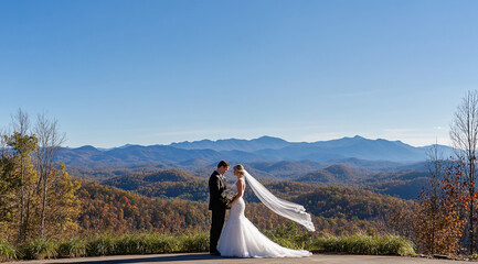 A bride and groom stand together on a mountaintop overlooking a stunning view of the Smoky Mountains. The wind blows through the bride's veil, adding a touch of magic to the moment