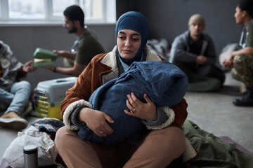 Portrait of distressed Middle Eastern woman with baby sitting on floor at refugee shelter and looking away, copy space