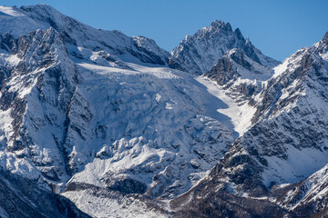 Obraz premium Snowy peaks and glaciers cover giant rocks. Pine and spruce forests grow at foot of mountains. which Majestic mountains against blue sky. Sunny day. Autumn 2024. Karachay-Cherkessia. Dombay.