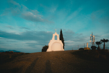 Serene white chapel with cross on a hilltop at sunset, surrounded by a vibrant blue sky and distant cypress