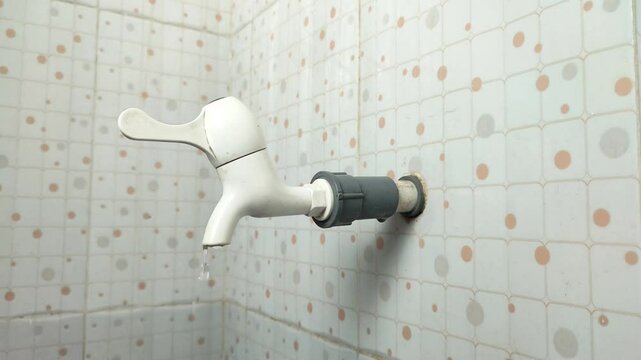 A hand turning off a white faucet on a tiled bathroom wall, with a small stream of water flowing out.
