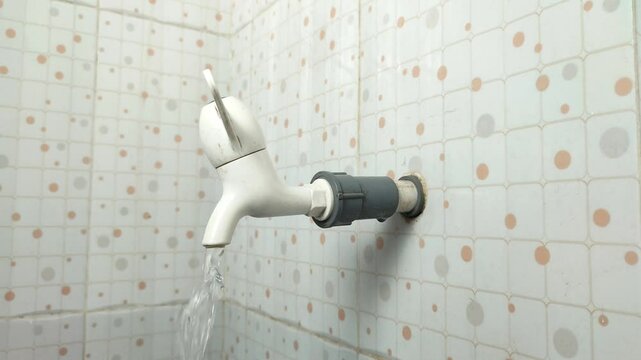 A hand turning on a white faucet on a tiled bathroom wall, with a small stream of water flowing out.