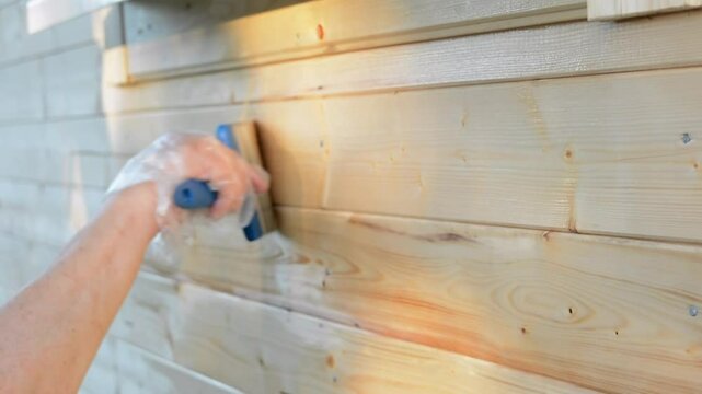 A hand wearing a protective plastic glove applies wood stain with a paintbrush to a wooden wall, focusing on even coverage and weather protection outdoors