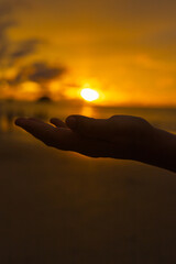 outstretched hand is silhouetted against a vibrant sunset over the ocean, with the sun descending...