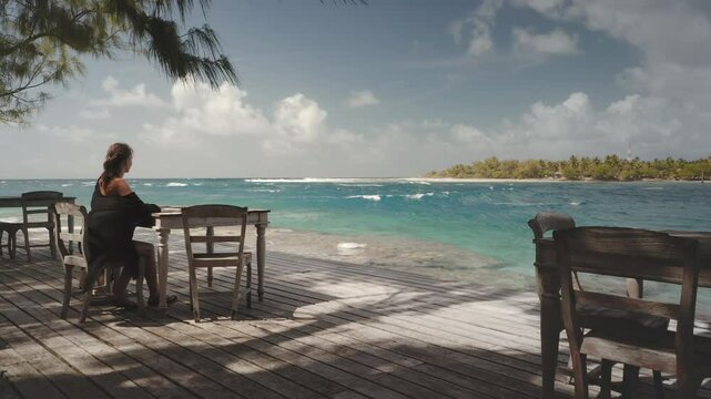 Young woman taking a break, sitting at a wooden table, enjoying the view of the turquoise ocean and crashing waves in a tropical paradise