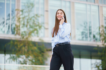 Fototapeta premium Young Woman in a Light Blue Shirt Talks on Her Phone While Walking Near a Modern Office Building in Spring